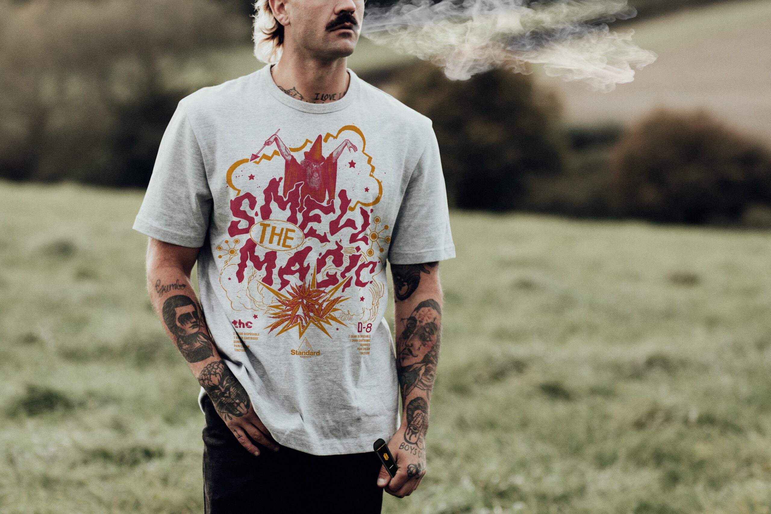 A man vaping, with tattoos on both his arms, wearing a T-shirt with the phrase "Smell the Magic".