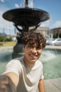 A man smiles as he takes a selfie in front of a beautiful fountain