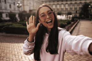 A smiling woman in glasses waves cheerfully at the camera.