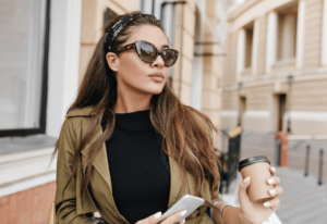 A woman wearing sunglasses and a trench coat, holding a coffee cup, stands stylishly on a city street.