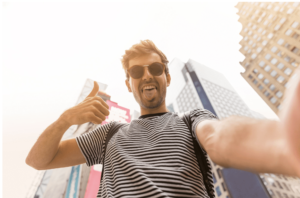 Man smiling while taking a selfie with his phone in a bustling city street.